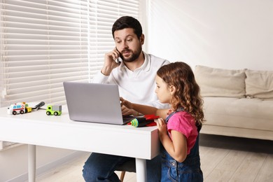 Photo of Naughty daughter disturbing her overwhelmed father while he talking on smartphone at table indoors