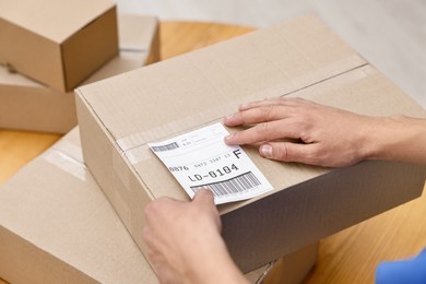 Photo of Man sticking shipping label with barcode on parcel at table indoors, closeup