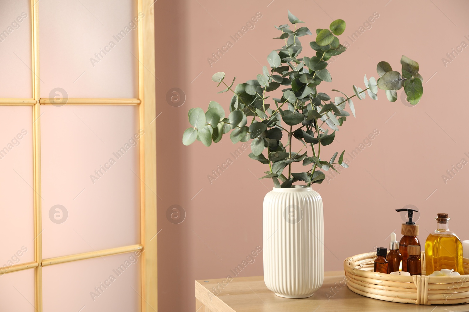 Photo of Eucalyptus branches in vase, cosmetic products and burning candles on wooden table indoors