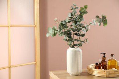 Photo of Eucalyptus branches in vase, cosmetic products and burning candles on wooden table indoors