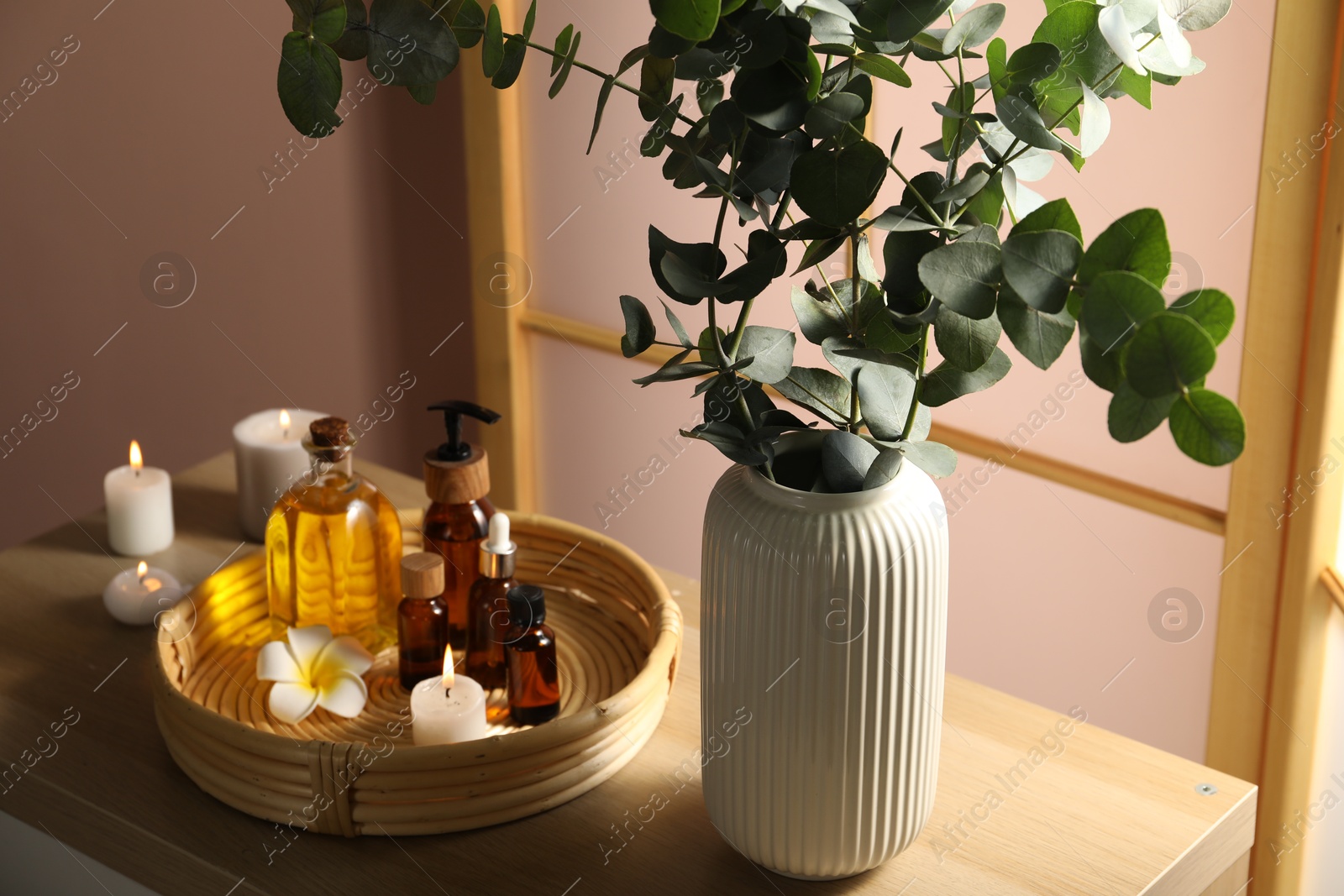 Photo of Eucalyptus branches in vase, cosmetic products and burning candles on wooden table indoors