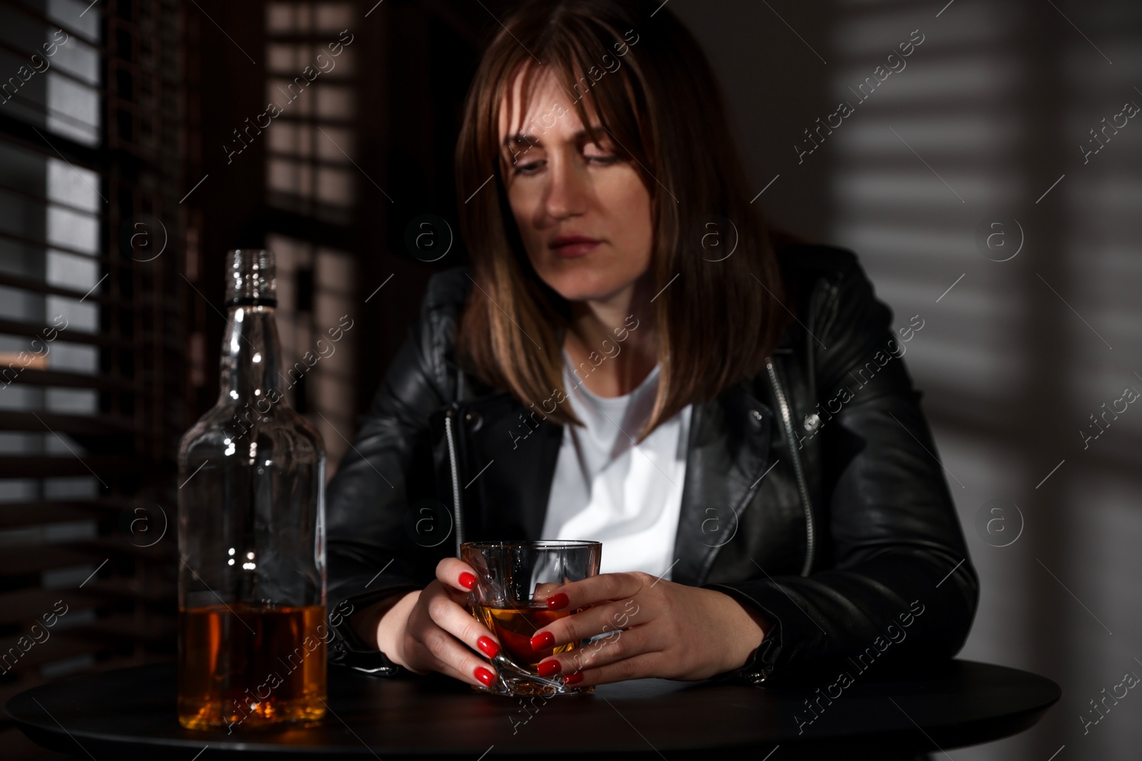 Photo of Alcohol addiction. Woman with glass of whiskey and bottle at table indoors