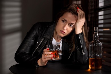 Photo of Alcohol addiction. Woman with glass of whiskey and bottle at table indoors