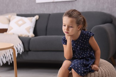 Photo of Resentful little girl sitting on pouf at home, space for text