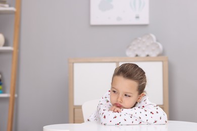 Photo of Resentful little girl at white table indoors, space for text