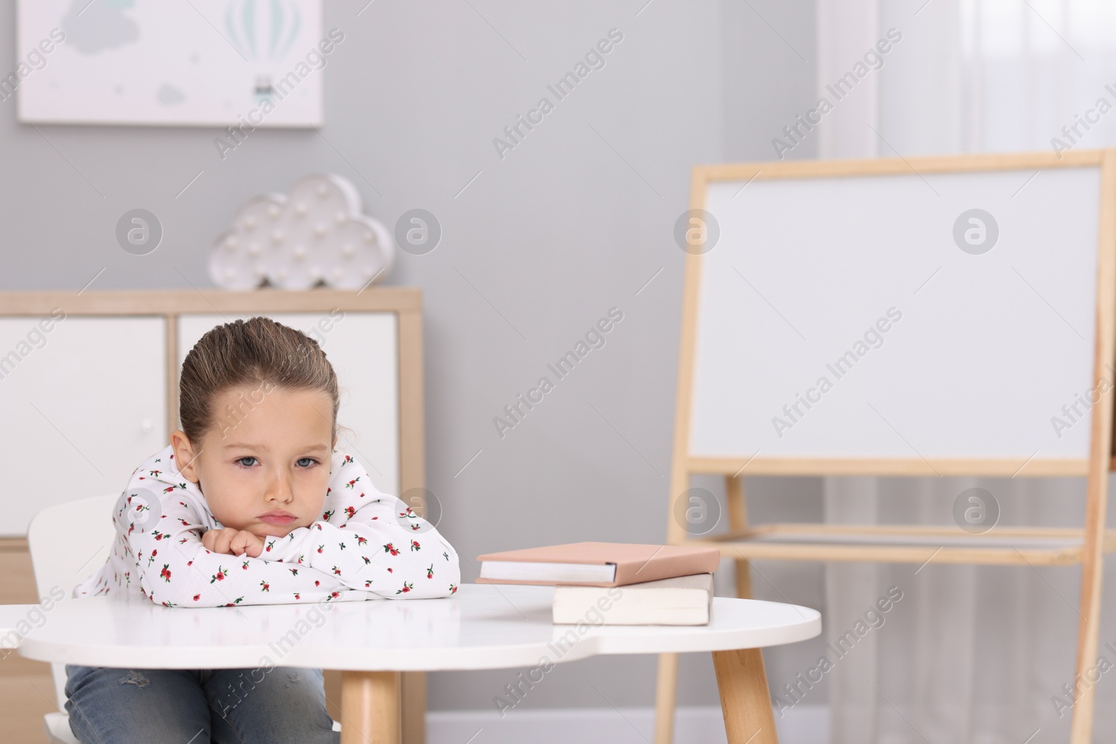 Photo of Resentful little girl at white table indoors, space for text