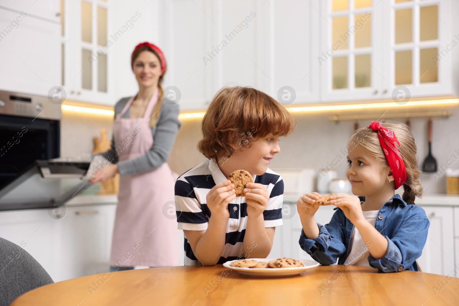 Photo of Happy kids eating tasty cookies while their mother baking in kitchen, selective focus