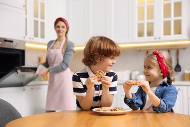 Photo of Happy kids eating tasty cookies while their mother baking in kitchen, selective focus