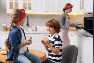 Photo of Happy kids eating tasty cookies while their mother baking in kitchen, selective focus