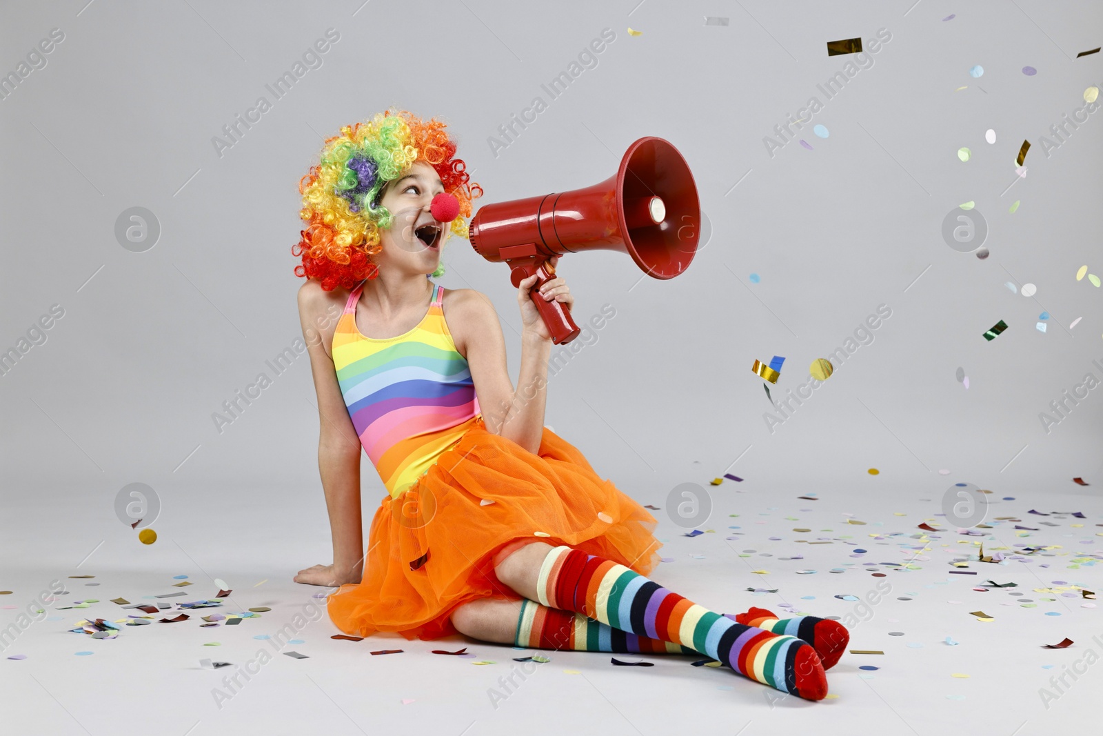 Photo of Girl dressed like clown shouting in megaphone under flying confetti on grey background. Surprise party