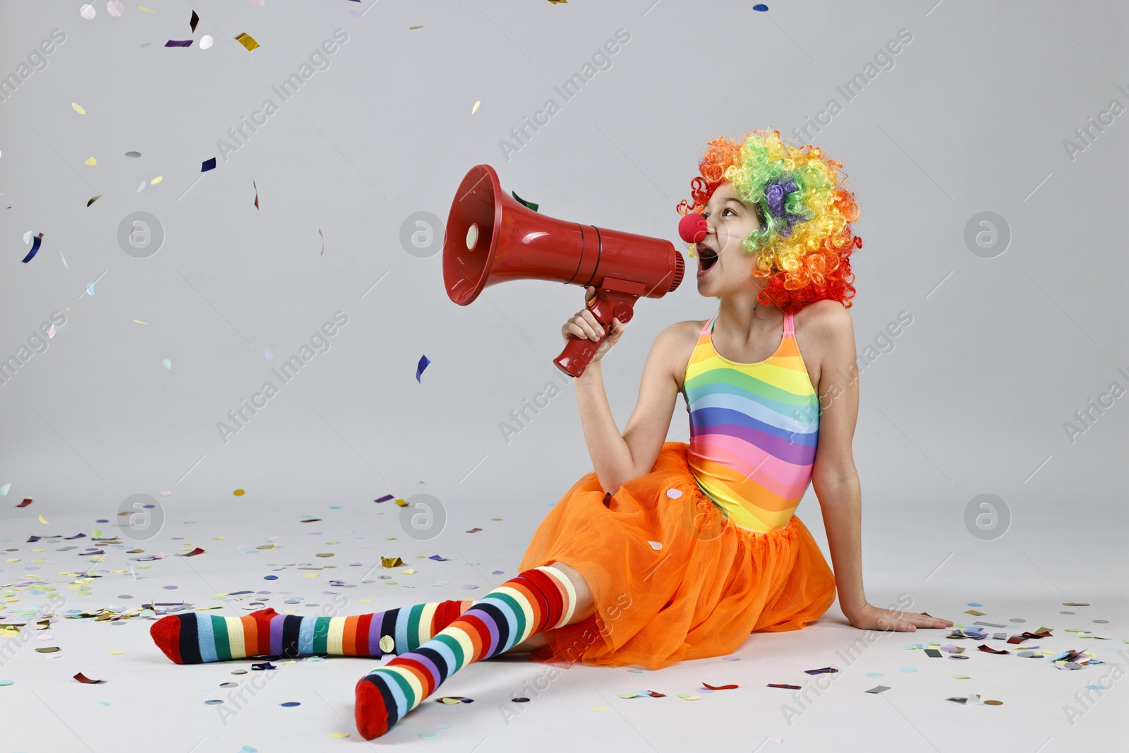 Photo of Girl dressed like clown shouting in megaphone under flying confetti on grey background. Surprise party