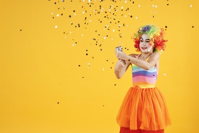 Photo of Happy girl dressed like clown with confetti popper on yellow background, space for text. Surprise party