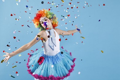 Photo of Happy girl dressed like clown under falling confetti on light blue background. Surprise party