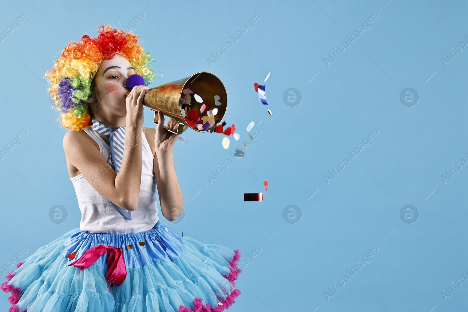 Photo of Happy girl dressed like clown with megaphone and confetti on light blue background, space for text. Surprise party