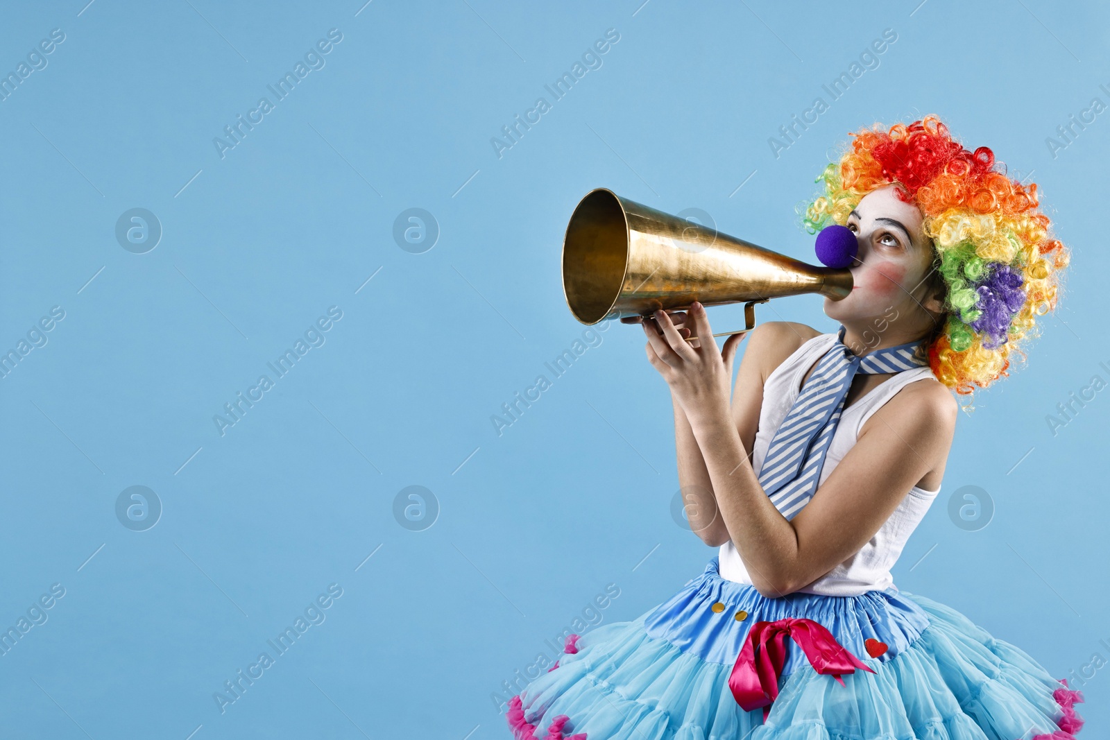 Photo of Happy girl dressed like clown with megaphone on light blue background, space for text. Surprise party