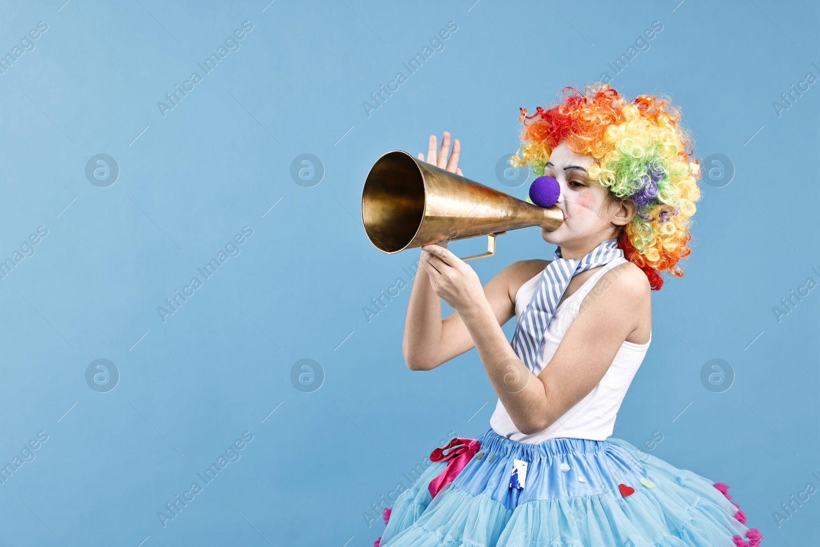 Photo of Happy little girl dressed like clown with megaphone on light blue background, space for text. Surprise party