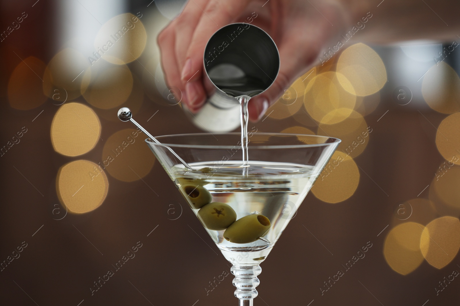 Photo of Woman making tasty martini cocktail with olives on blurred background, closeup. Bokeh effect