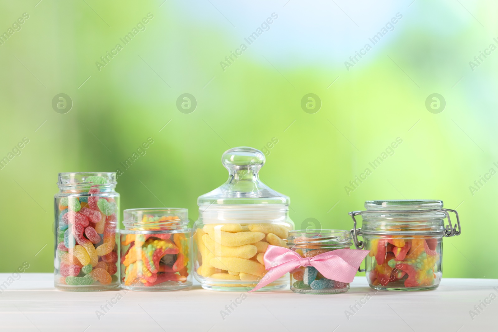 Photo of Tasty gummy candies in jars on white wooden table against blurred green background