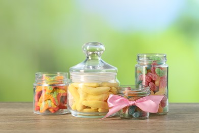 Photo of Tasty gummy candies in jars on wooden table against blurred green background