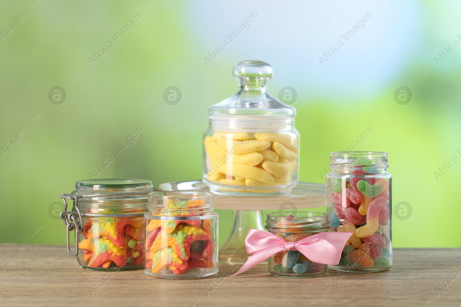 Photo of Tasty gummy candies in jars on wooden table against blurred green background