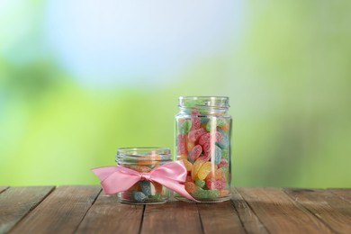 Photo of Tasty gummy candies in jars on wooden table against blurred green background