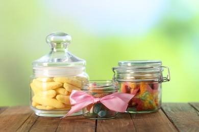 Photo of Tasty gummy candies in jars on wooden table against blurred green background