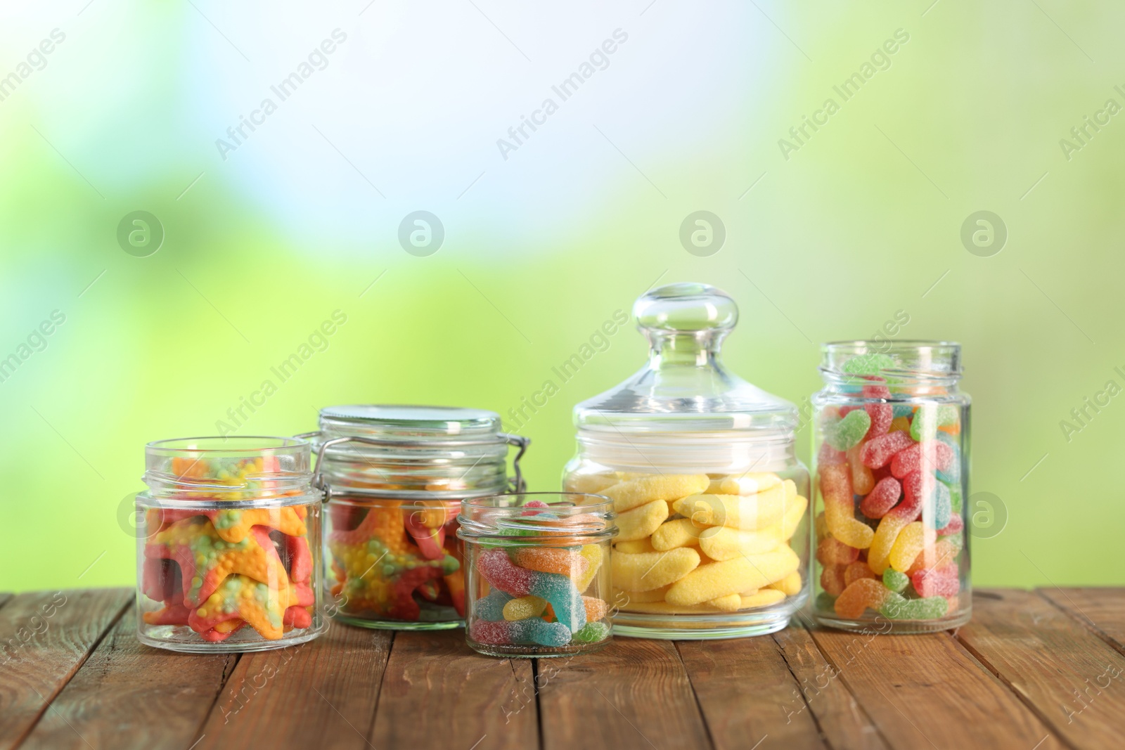 Photo of Tasty gummy candies in jars on wooden table against blurred green background