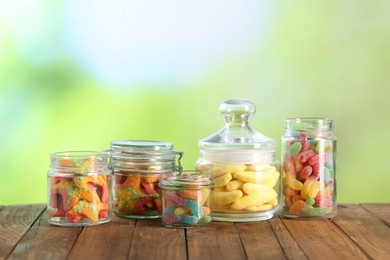 Photo of Tasty gummy candies in jars on wooden table against blurred green background