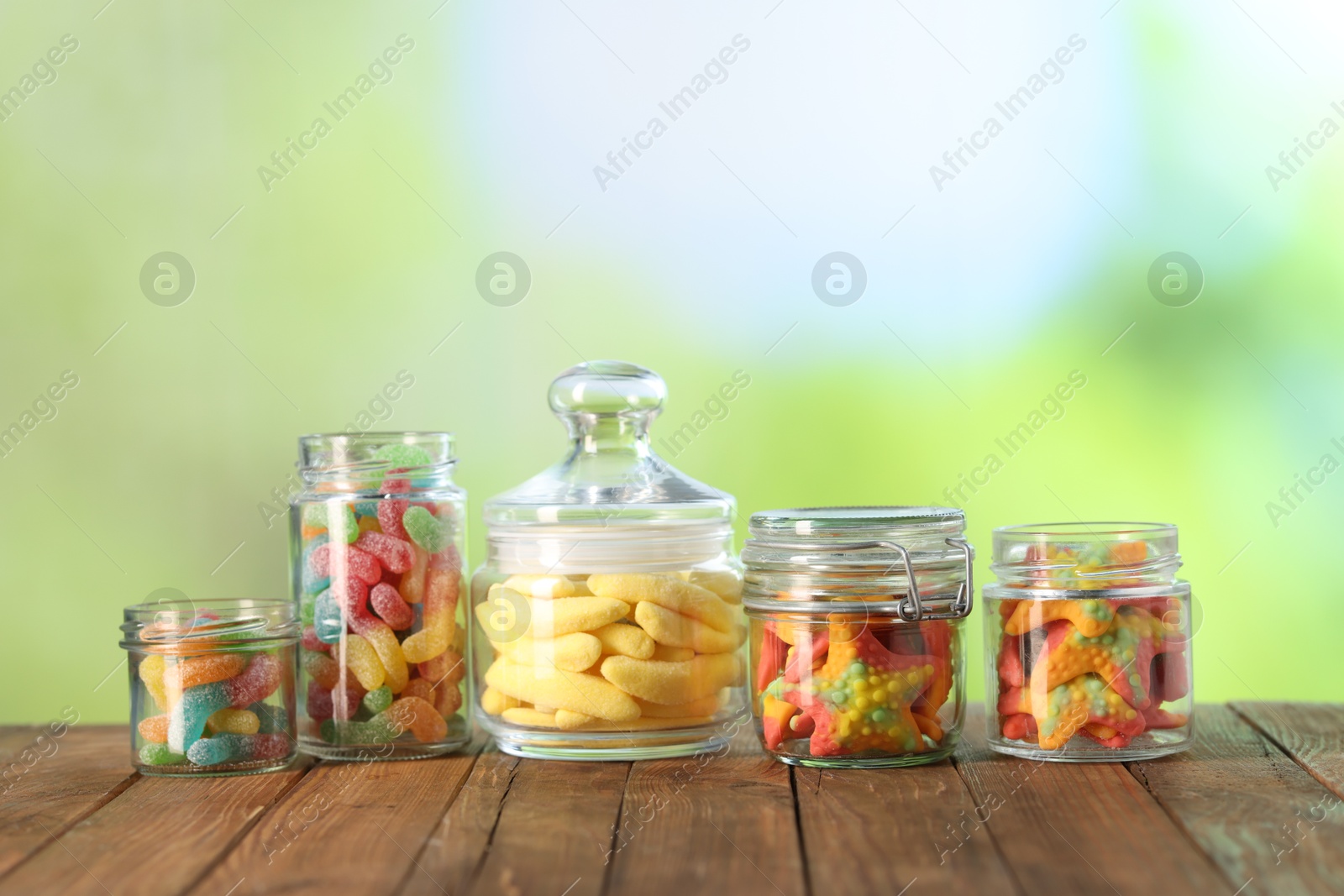 Photo of Tasty gummy candies in jars on wooden table against blurred green background