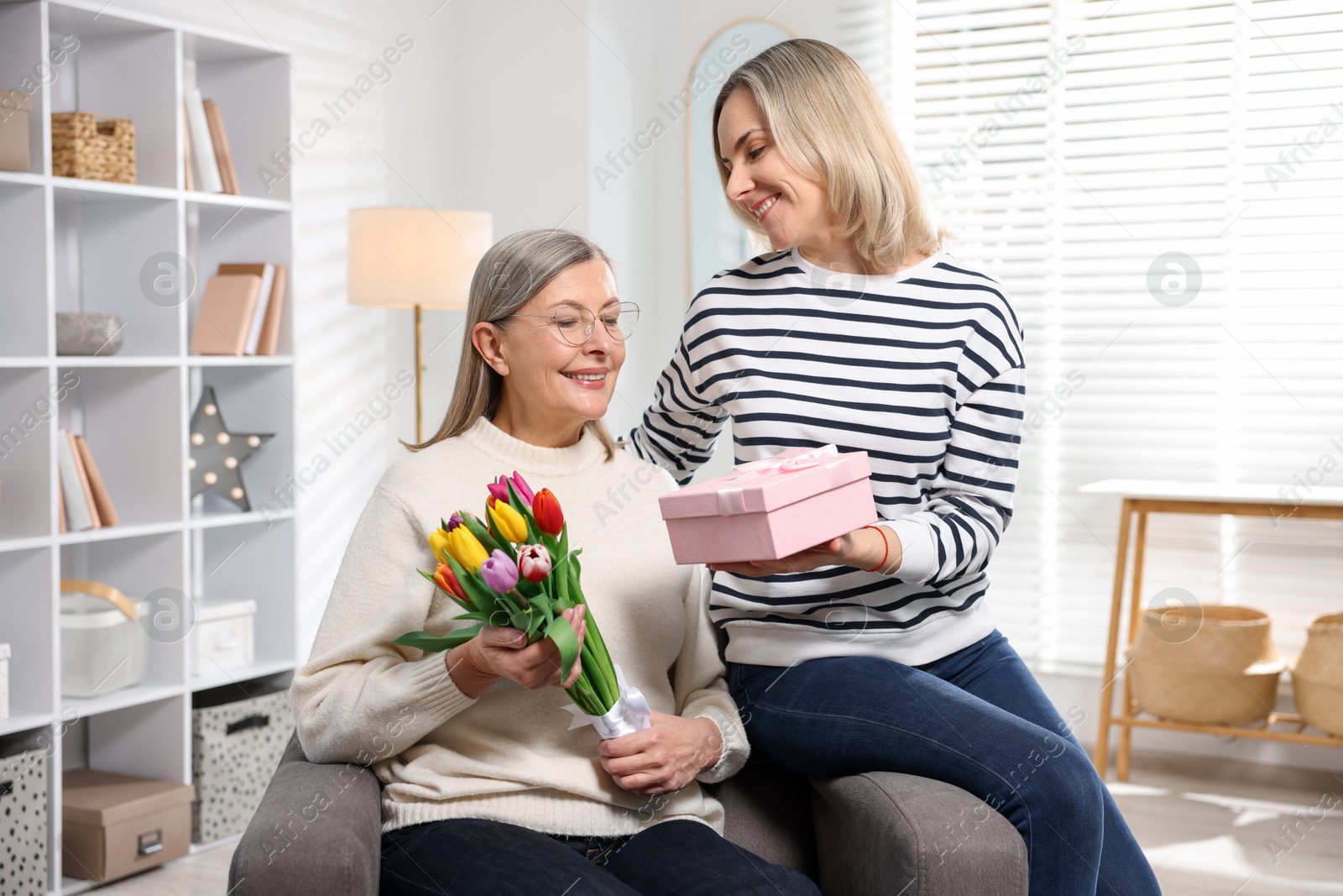 Photo of Smiling daughter congratulating her mom with bouquet of tulips and gift at home. Happy Mother's Day