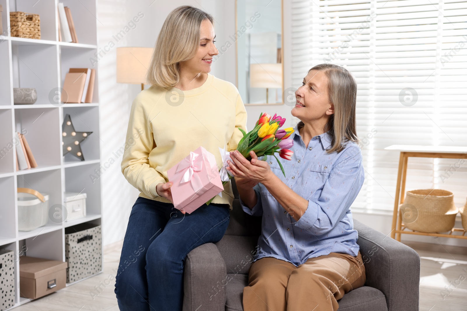 Photo of Smiling daughter congratulating her mom with bouquet of tulips and gift on armchair at home. Happy Mother's Day