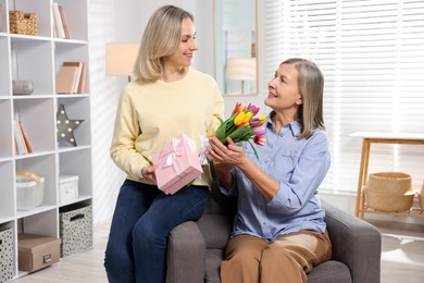 Photo of Smiling daughter congratulating her mom with bouquet of tulips and gift on armchair at home. Happy Mother's Day