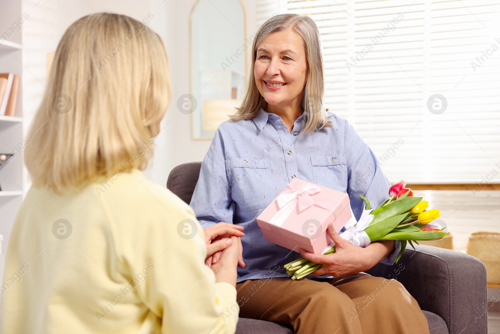 Photo of Smiling mom receiving bouquet of tulips and gift from her daughter on armchair at home. Happy Mother's Day