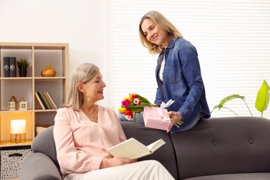 Photo of Smiling daughter congratulating her mom with bouquet of tulips and gift on sofa at home. Happy Mother's Day