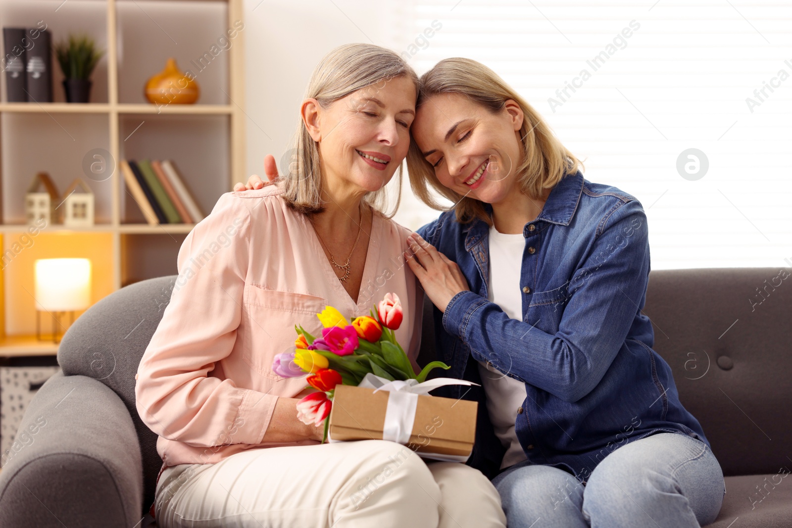 Photo of Smiling woman with bouquet of tulips, gift and her daughter on sofa at home. Happy Mother's Day