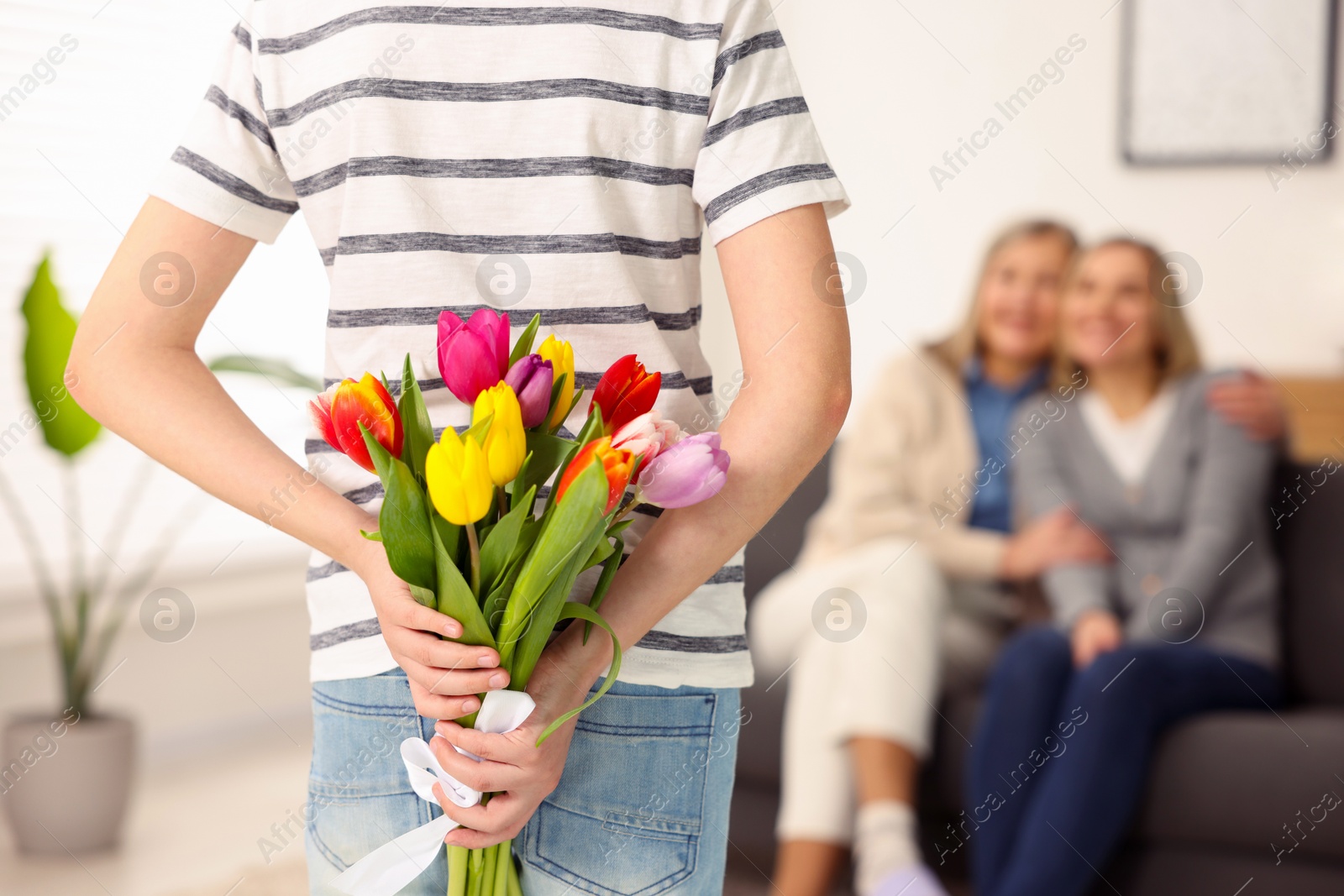 Photo of Boy hiding bouquet of tulips behind back for his parents at home, selective focus. Happy Mother's Day