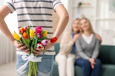 Photo of Boy hiding bouquet of tulips behind back for his parents at home, selective focus. Happy Mother's Day