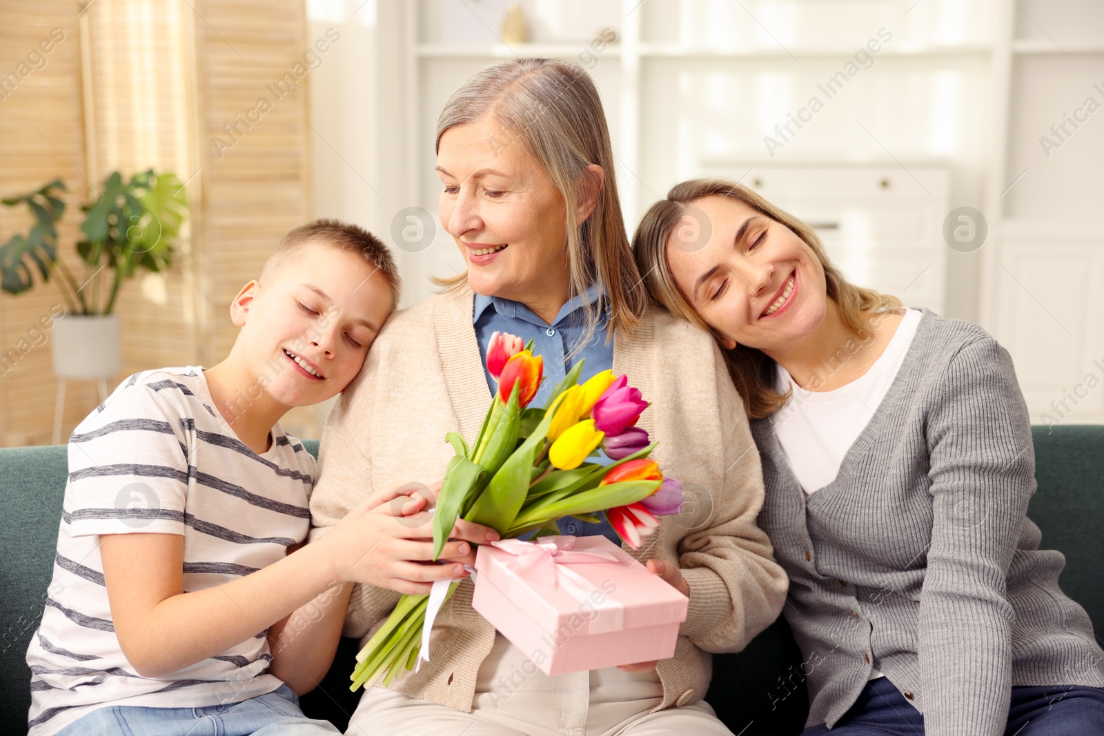 Photo of Smiling woman with bouquet of tulips, gift box and her relatives on sofa at home. Happy Mother's Day