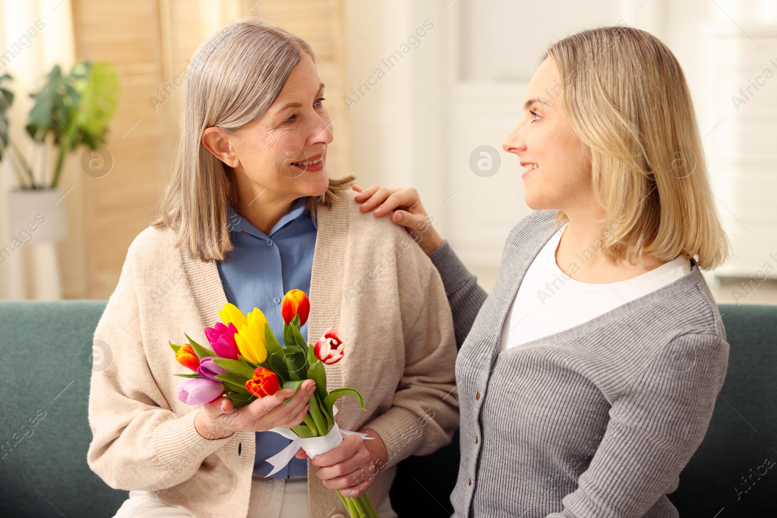 Photo of Smiling woman with bouquet of tulips and her daughter on sofa at home. Happy Mother's Day