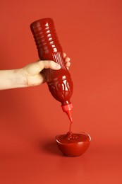 Photo of Woman squeezing ketchup from bottle into bowl on red background, closeup