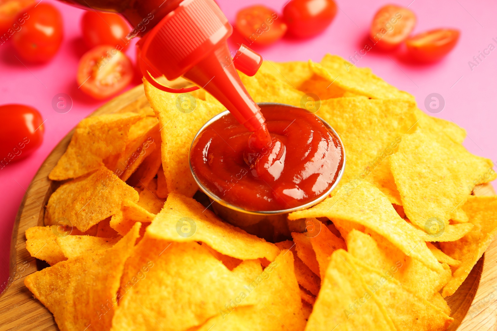 Photo of Pouring tasty ketchup in bowl with nachos on pink background, closeup