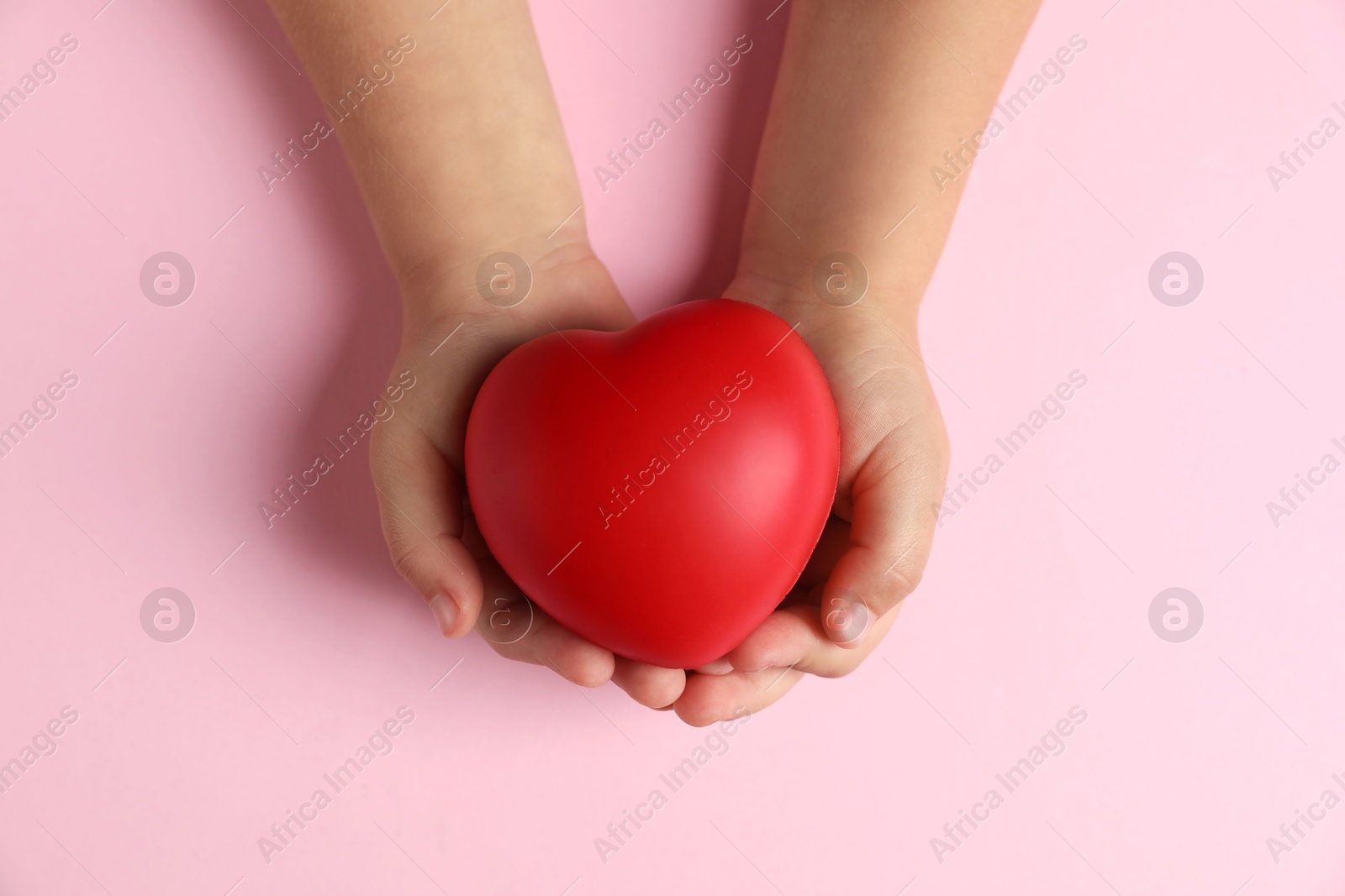 Photo of Child holding decorative red heart on light pink background, top view