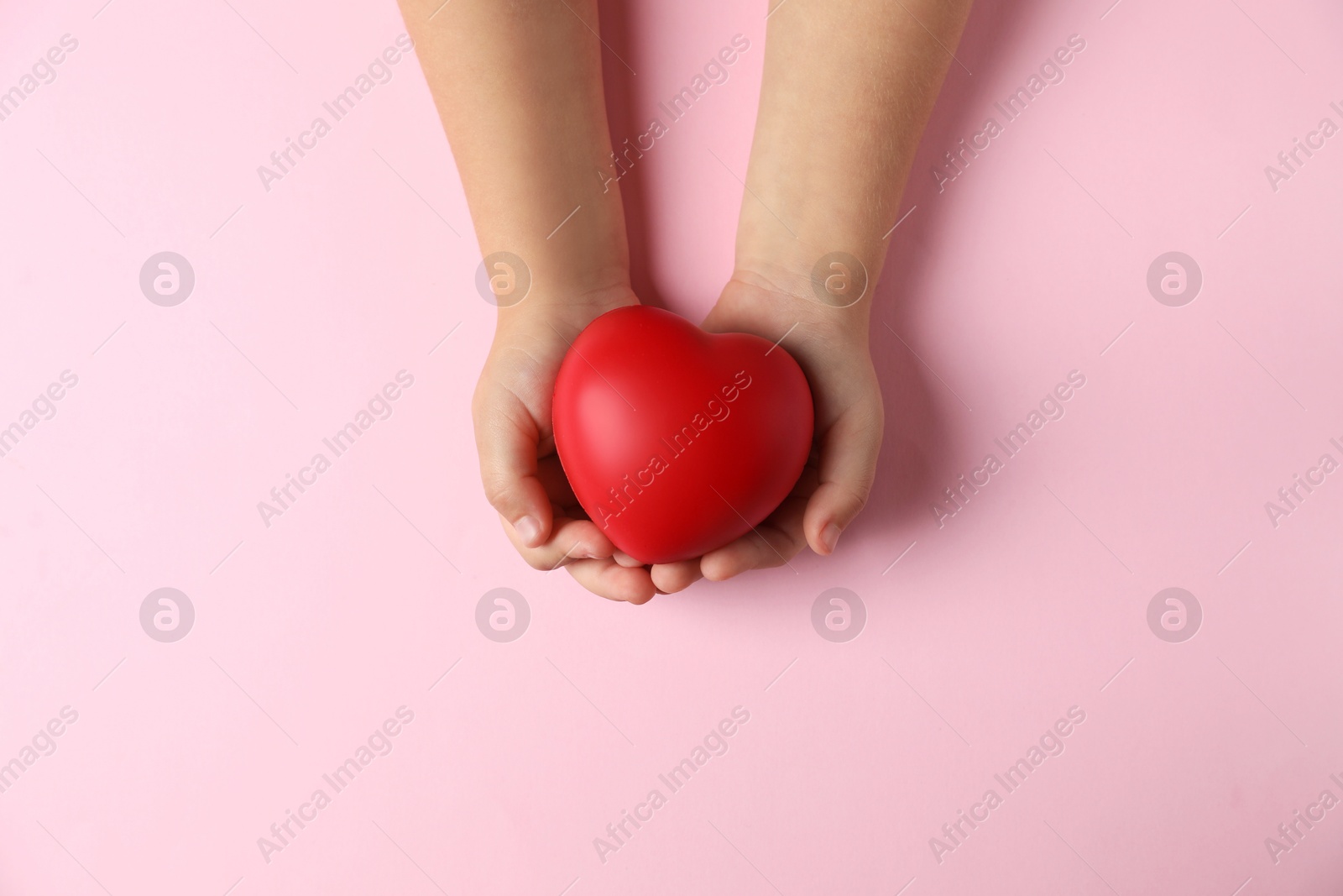 Photo of Child holding decorative red heart on light pink background, top view