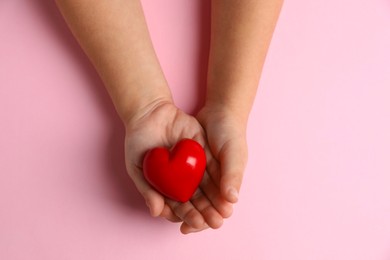 Photo of Child holding decorative red heart on light pink background, top view