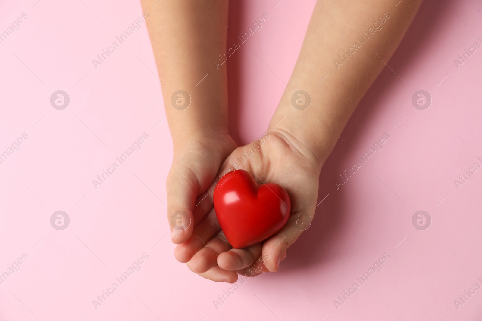 Photo of Child holding decorative red heart on light pink background, top view