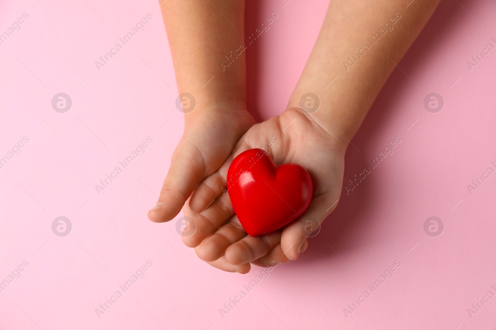 Photo of Child holding decorative red heart on light pink background, top view