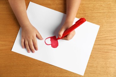 Photo of Little child drawing heart with felt tip pen at wooden table, top view