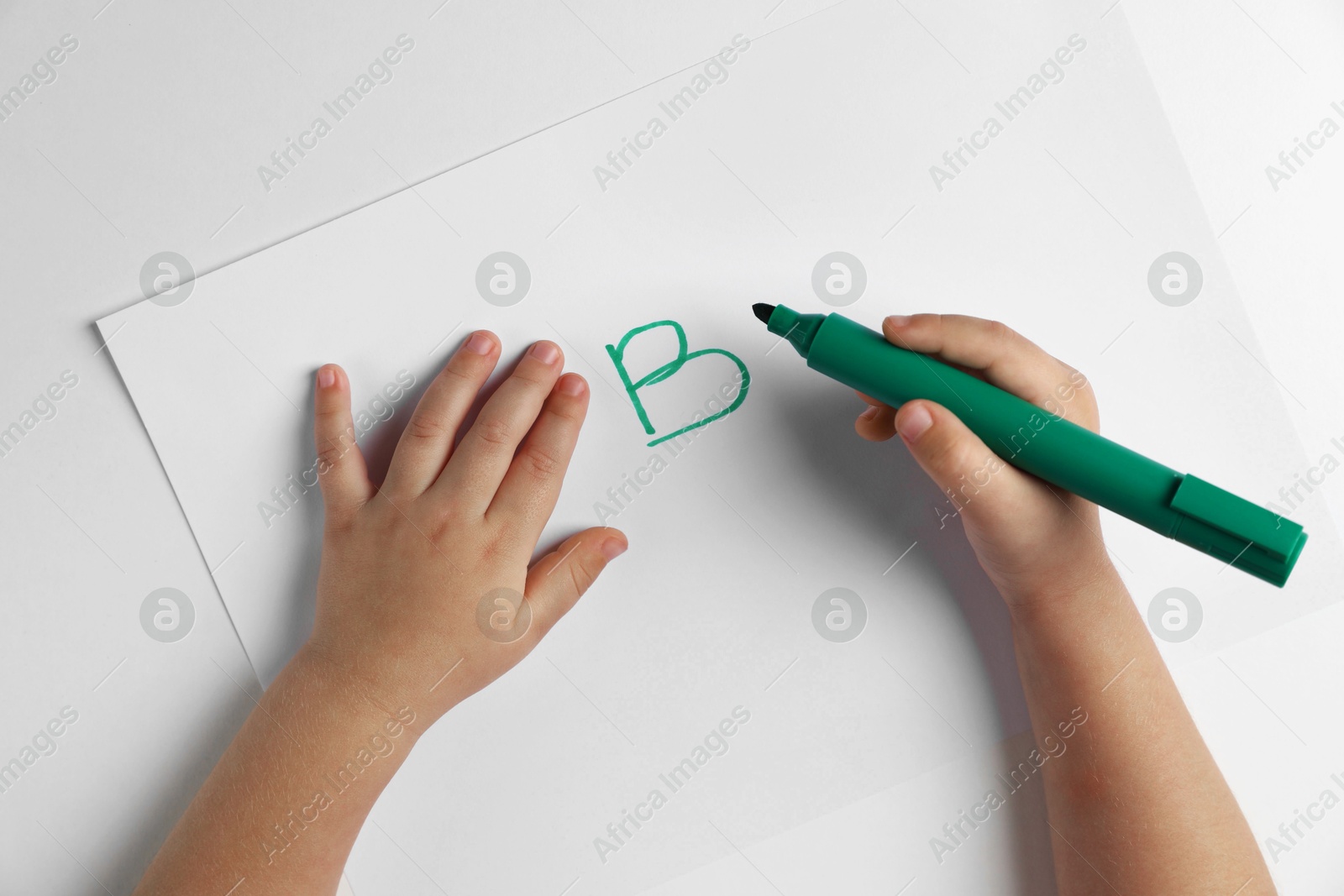 Photo of Little child writing letter B on paper sheet at light table, top view