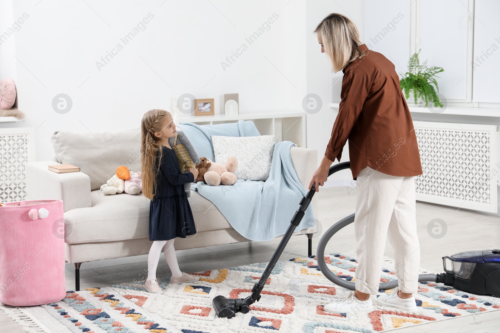 Photo of Little helper. Girl putting away toys while her mother vacuuming at home