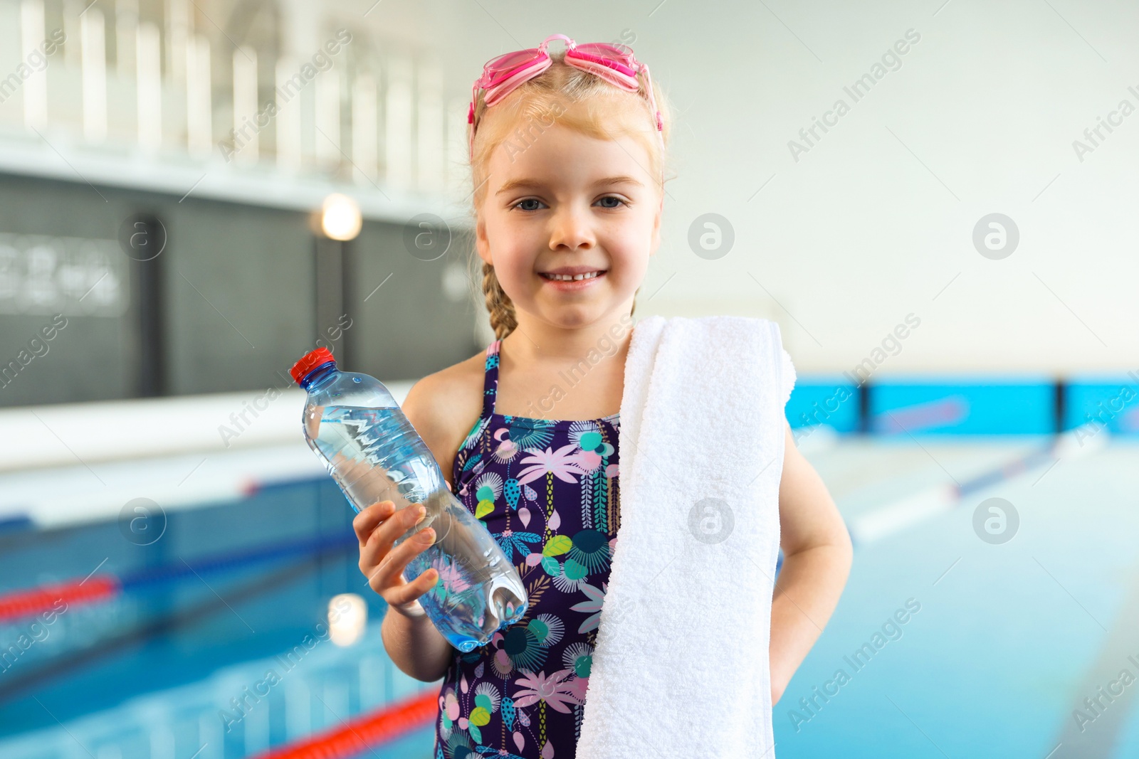 Photo of Little girl with goggles, towel and water near swimming pool indoors